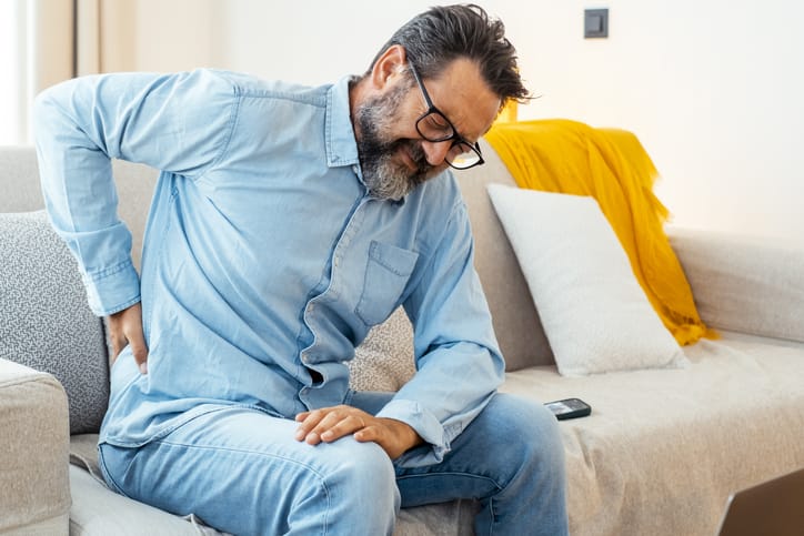 One man massaging his lower back after using laptop sitting on sofa for bad posture sitting. People unhealthy habits workplace at home. Mature male massage his body with painful expression on face