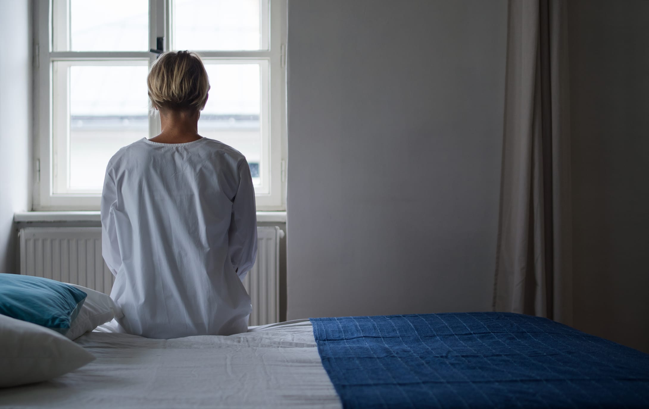Woman in hospital gown sitting on bed looking out window