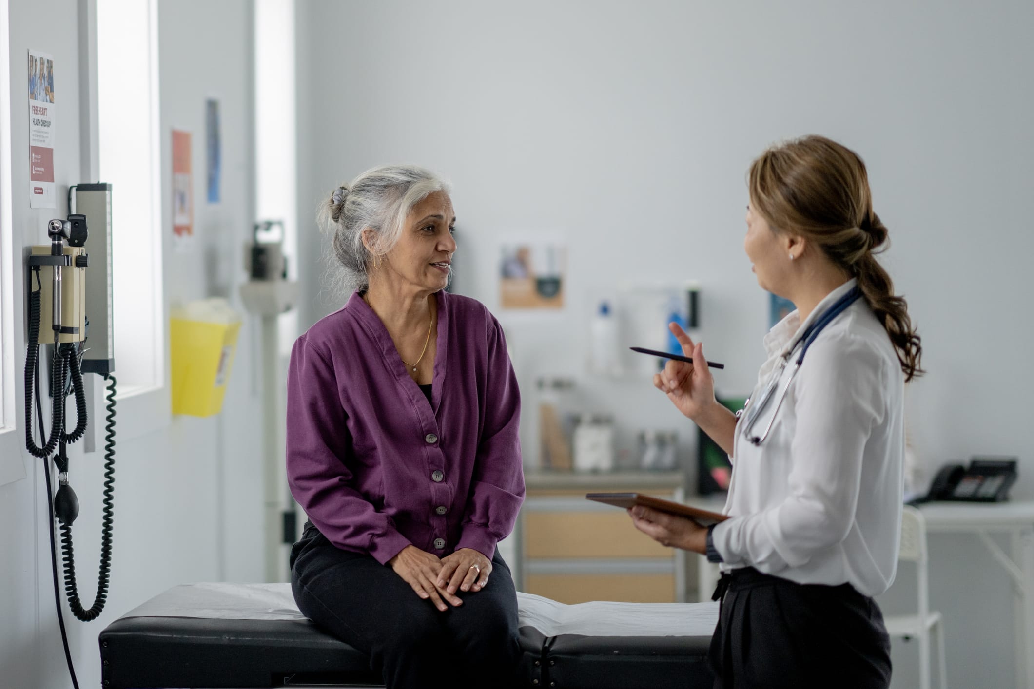 A senior woman of mixed race, sits up on an exam table across from her female doctor of Asian decent, as they discuss her health concerns.